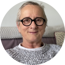 Photo of Booney Smith. Booney is a white woman with short grey hair and black glasses. She's sitting on a cream sofa and wearing a grey and white jumper over a white shirt.